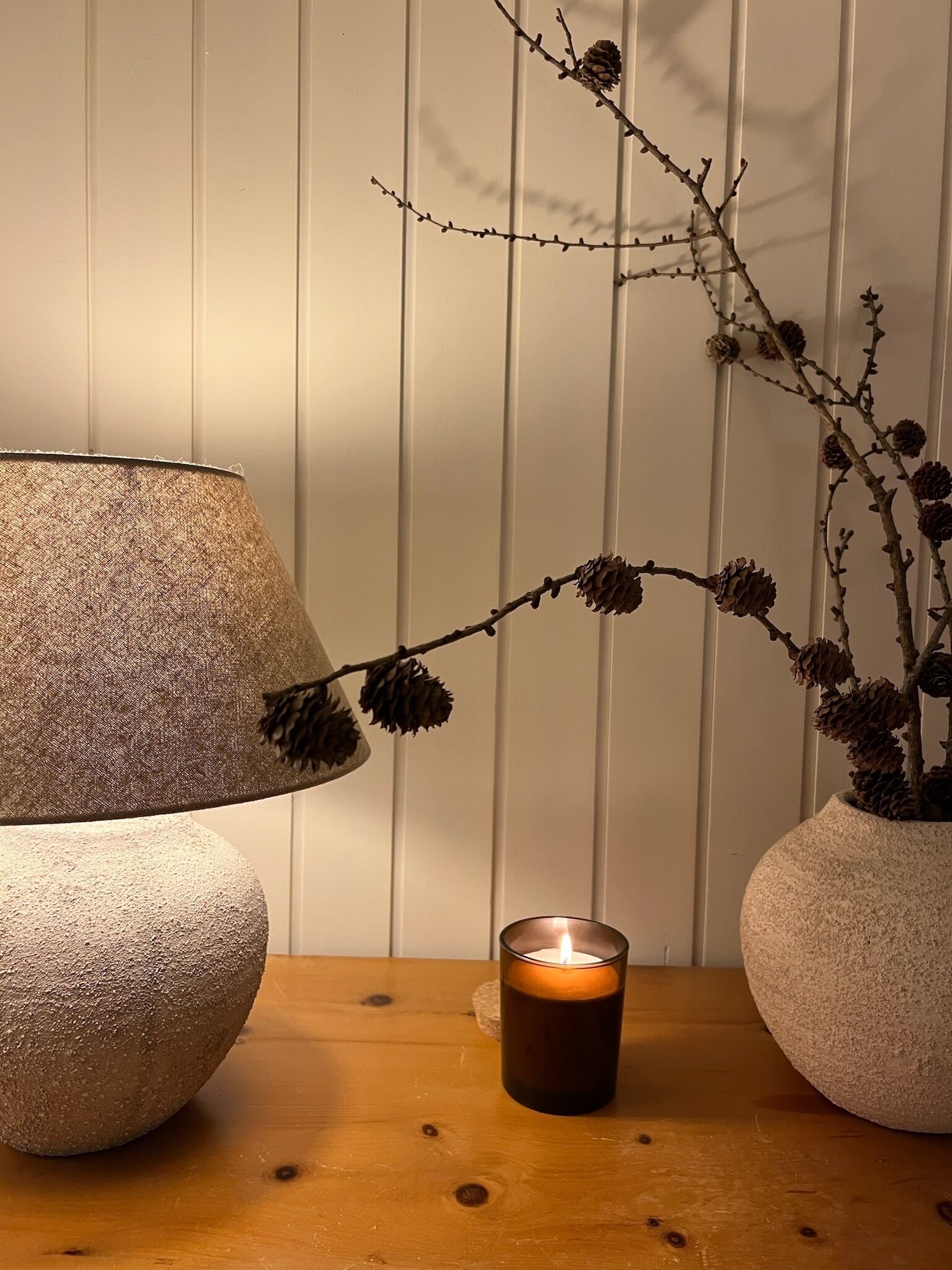 A lamp, a candle, and a branch with pine cones on a wooden table against a white wall.