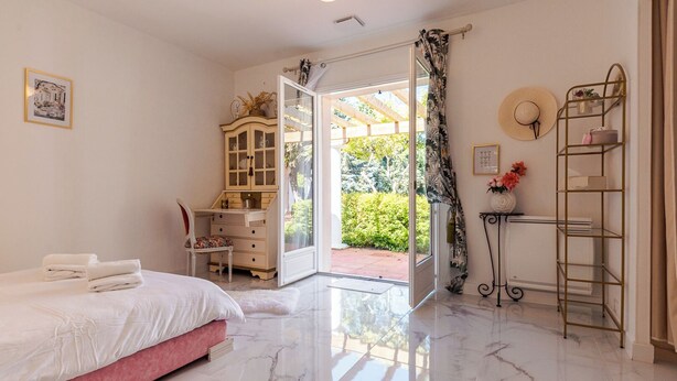 Une chambre à coucher comprenant un lit, un bureau, une armoire et donnant sur un jardin grâce à une porte ouverte.