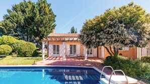 Une piscine à l’eau d’un bleu cristallin, un patio couvert avec des meubles d’extérieur, et une maison avec un toit en tuiles et des portes blanches.