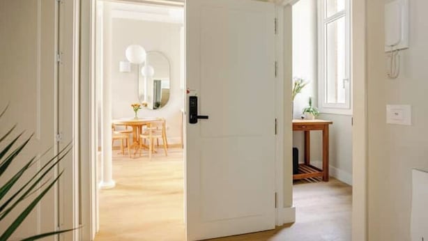 Um corredor com uma porta branca que dá para um quarto com piso de madeira e uma pequena mesa com uma planta.