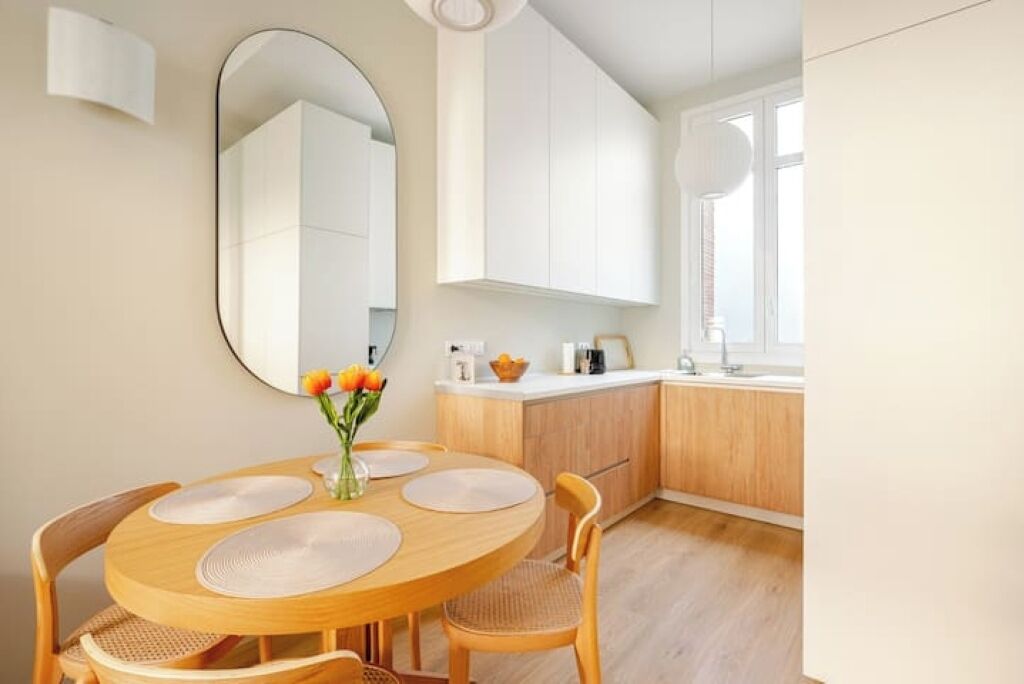 A modern kitchen with white cabinets, a round wooden dining table with placemats, and a vase with flowers.