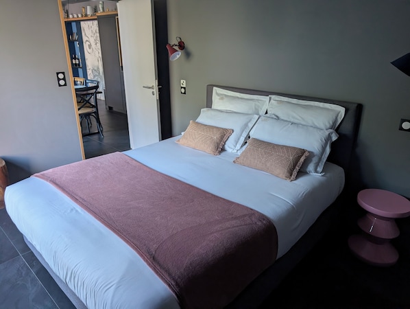 Ein ordentlich bezogenes Bett mit einer rosa und weißen Tagesdecke, zwei beigen Kissen und einem Kopfteil. Eine kleine, runde Tisch mit einem rosa Fuß ist rechts vom Bett zu sehen.
