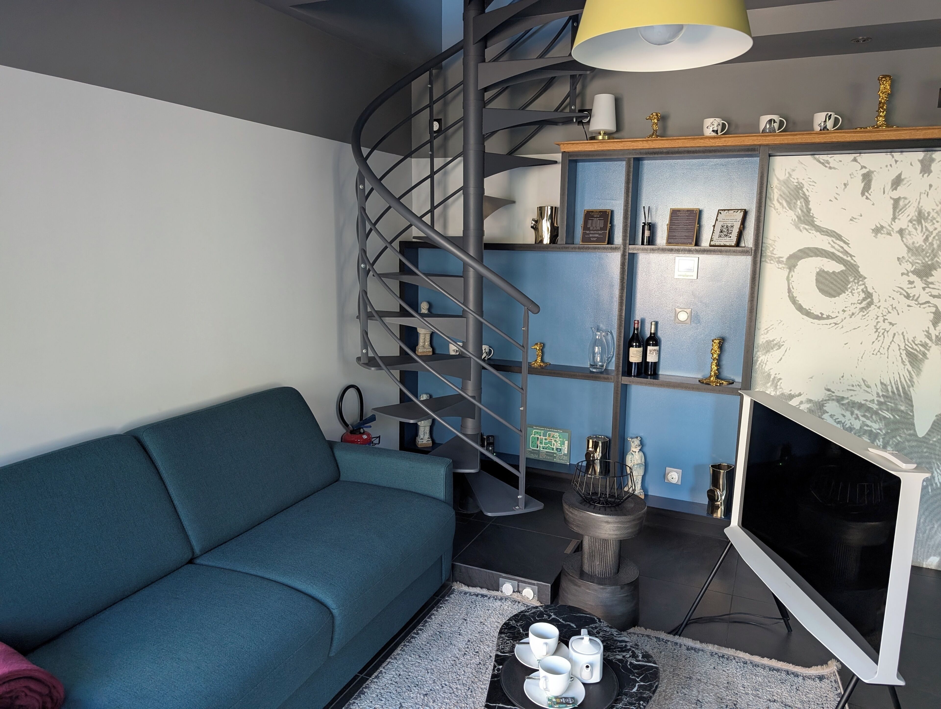 Un salon moderne avec un canapé bleu, un escalier en colimaçon, un téléviseur à écran plat et une étagère ornée d’objets décoratifs et de bouteilles de vin.