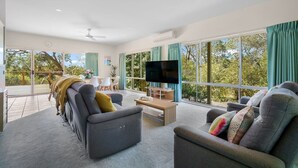 Ein modernes Wohnzimmer mit einem Flachbildfernseher, grauen Sofas, einem Couchtisch und großen Fenstern mit Blick auf Grünflächen.