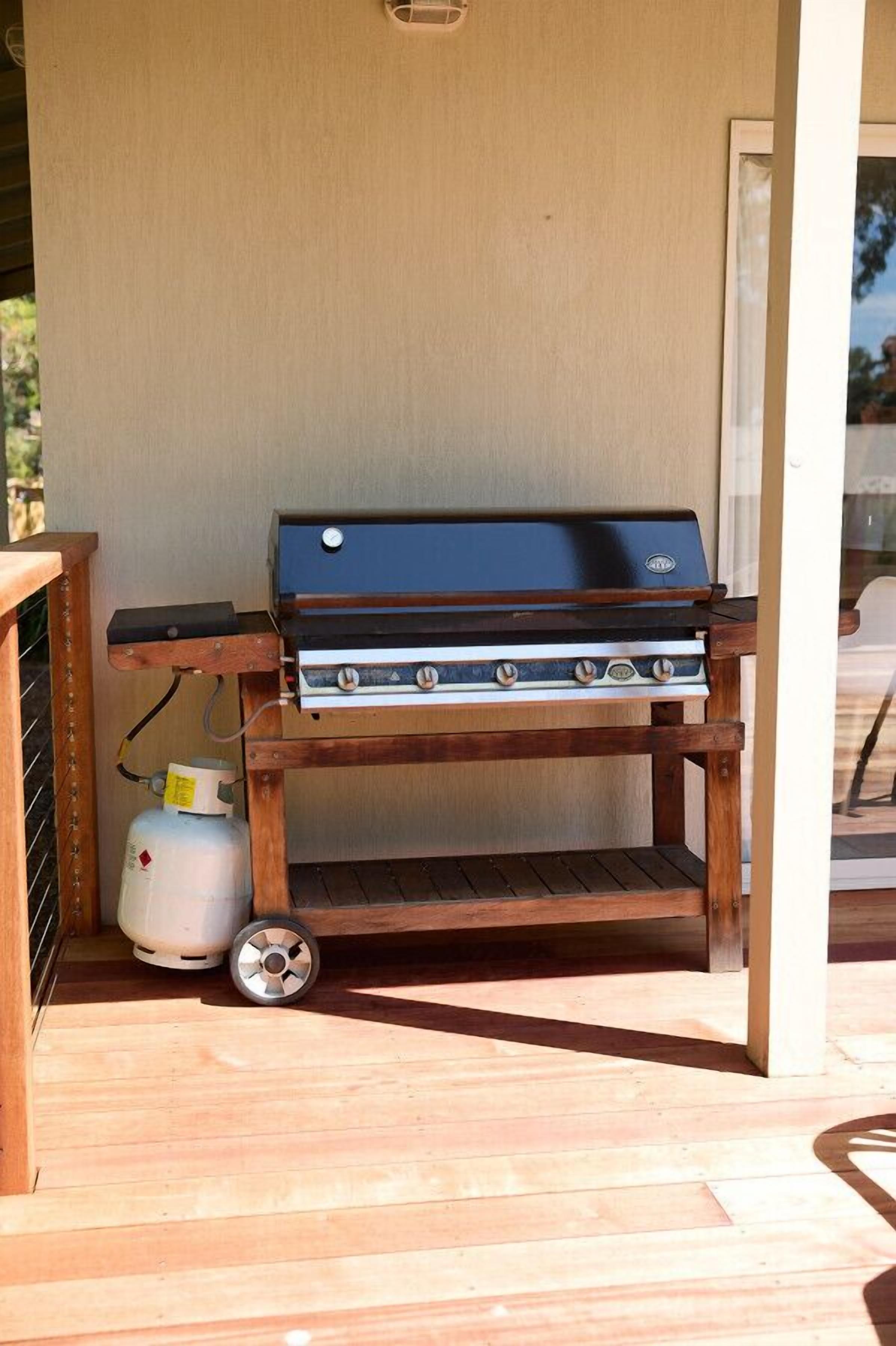 Ein tragbarer Grill mit Holzgestell und Rollen, ein Propangasbehälter und eine Glastür im Hintergrund.