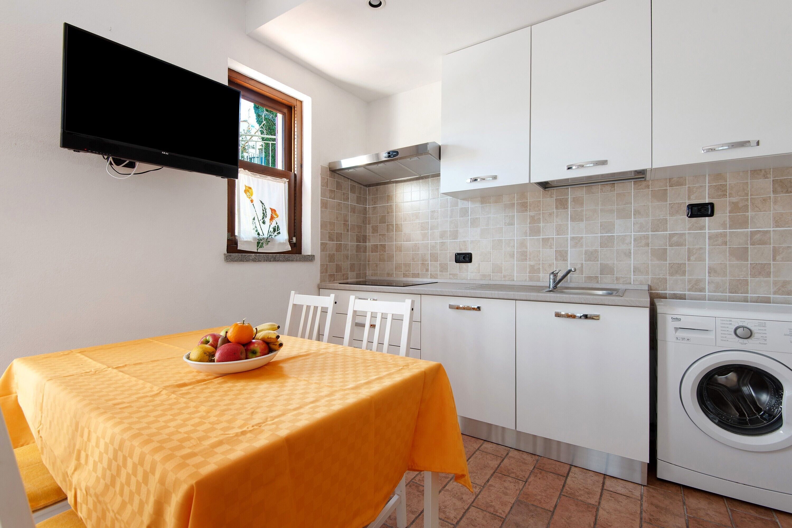 A kitchen with a washing machine, a dining table with a fruit bowl, white cabinets, and a flat-screen TV mounted on the wall.