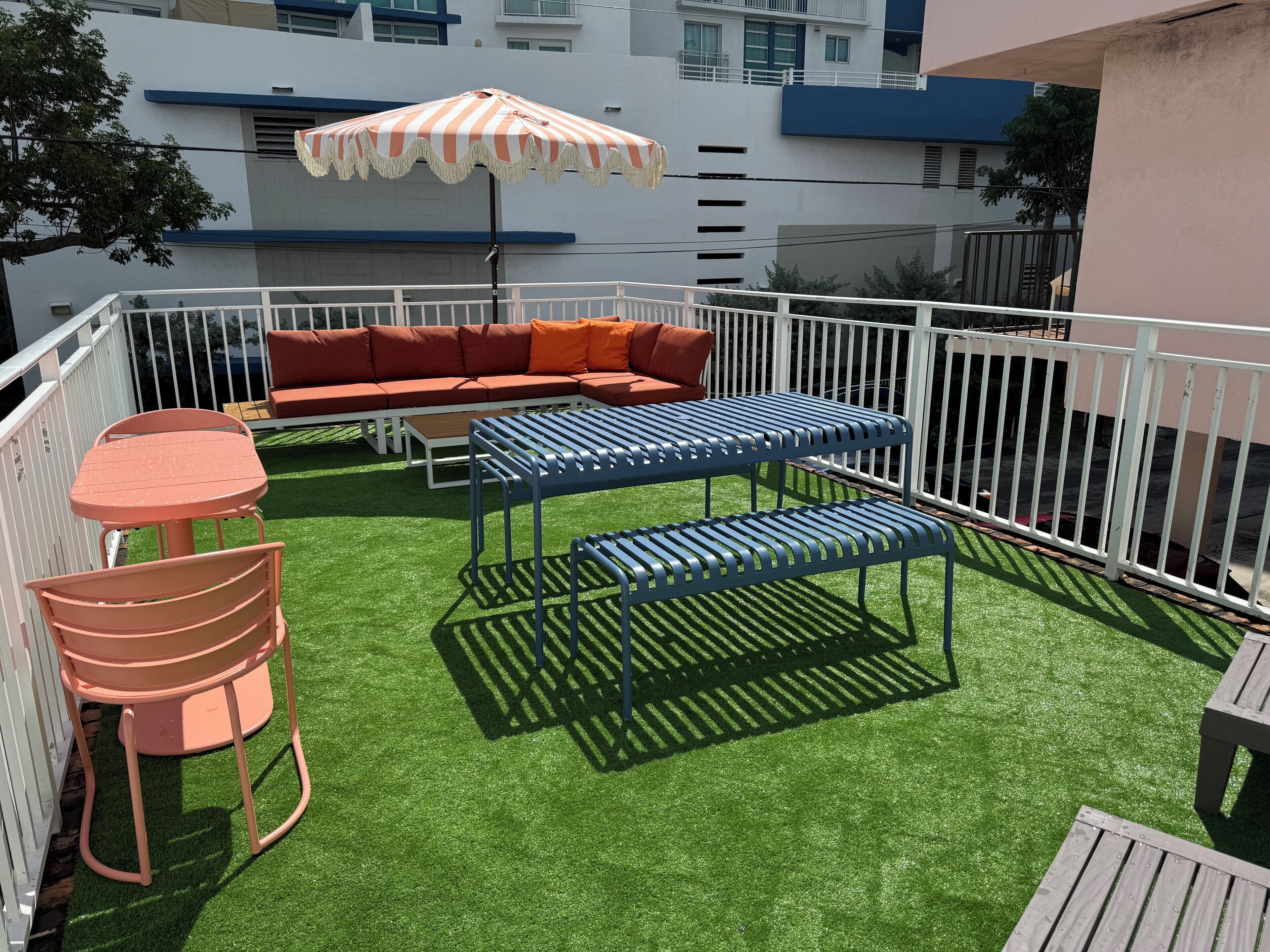 Eine Terrasse im Freien mit künstlichem Gras, einem gestreiften Sonnenschirm, einer Couch sowie Metalltischen und -stühlen.