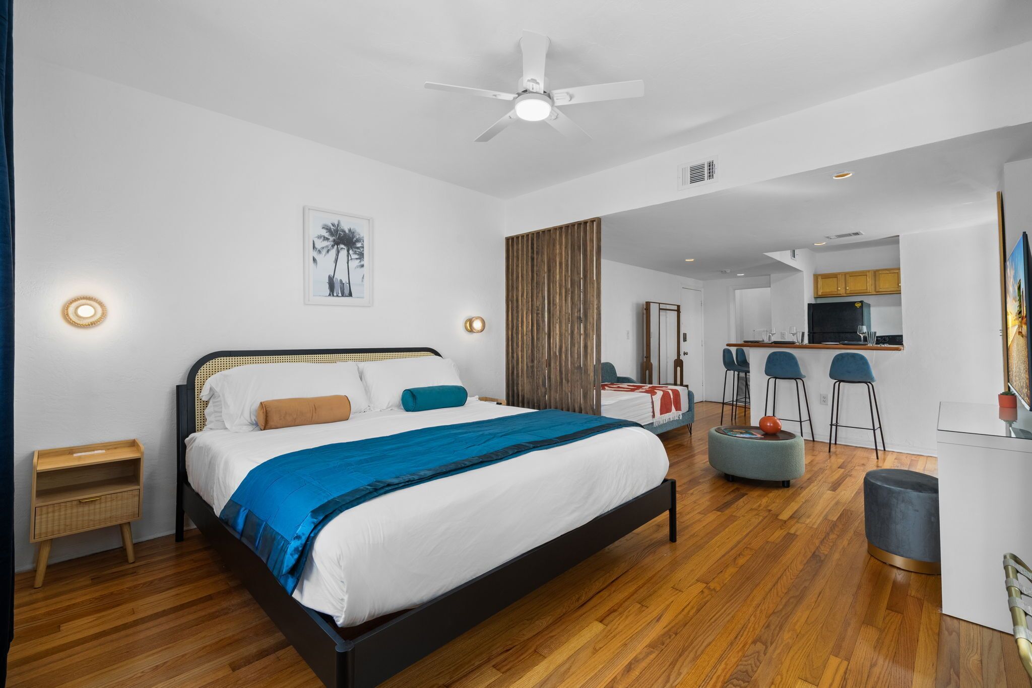 A modern hotel room with a large bed, a ceiling fan, a wooden floor, a kitchenette, and a dining area with bar stools.