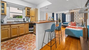A modern kitchen with wooden cabinets, a central island with bar stools, and a dining area with a sofa and curtains. - Blue Flamingo Suites (Miami Beach)
