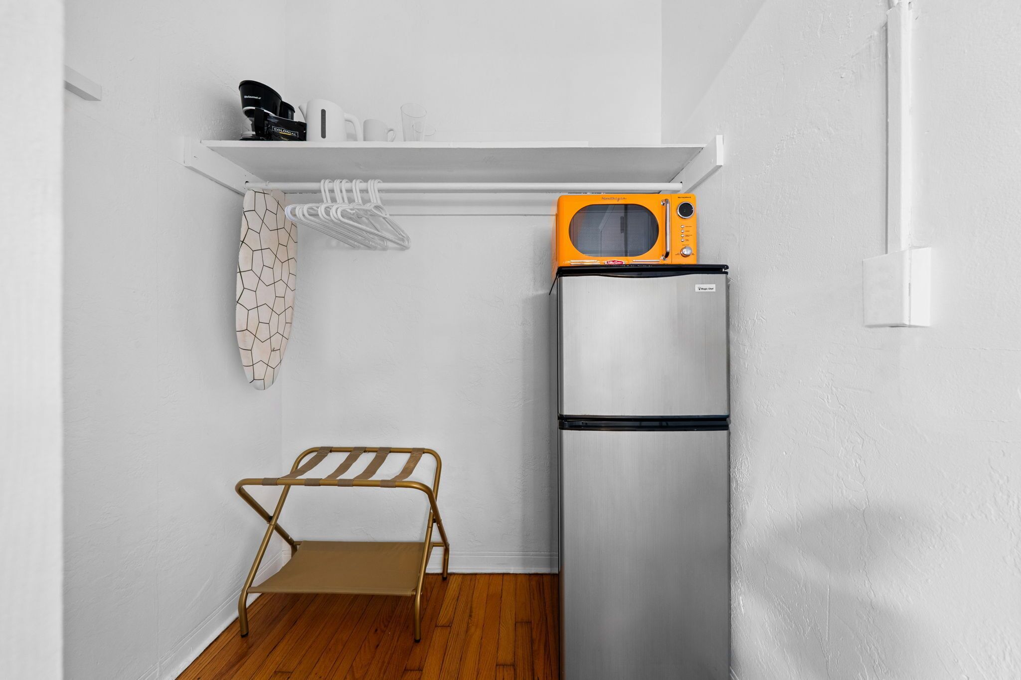 A compact kitchen with a stainless steel refrigerator, a microwave, and a small shelf with kitchen items.