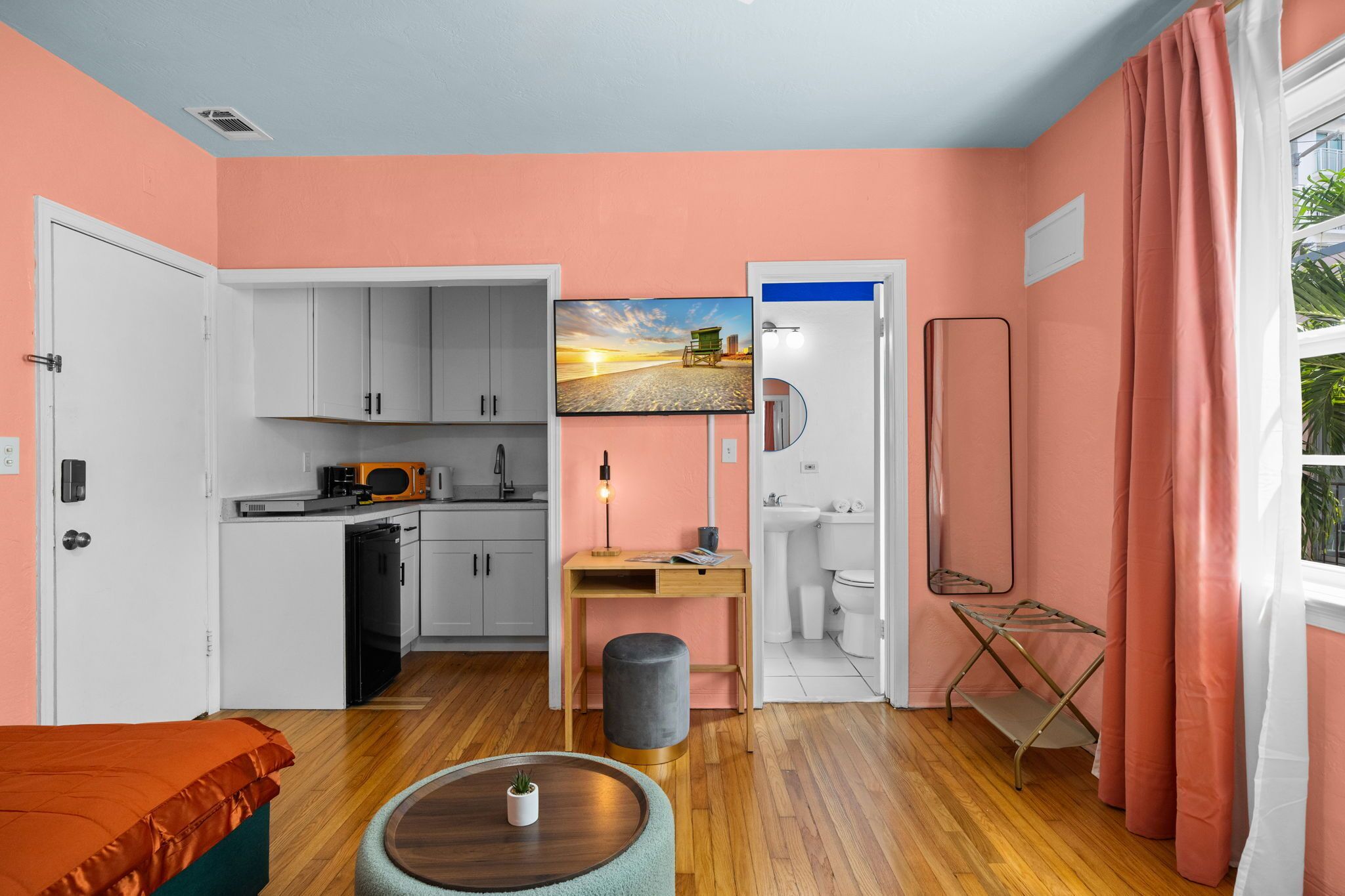 A compact room with a kitchenette, a bed, a small table with a plant, and a painting on the wall.