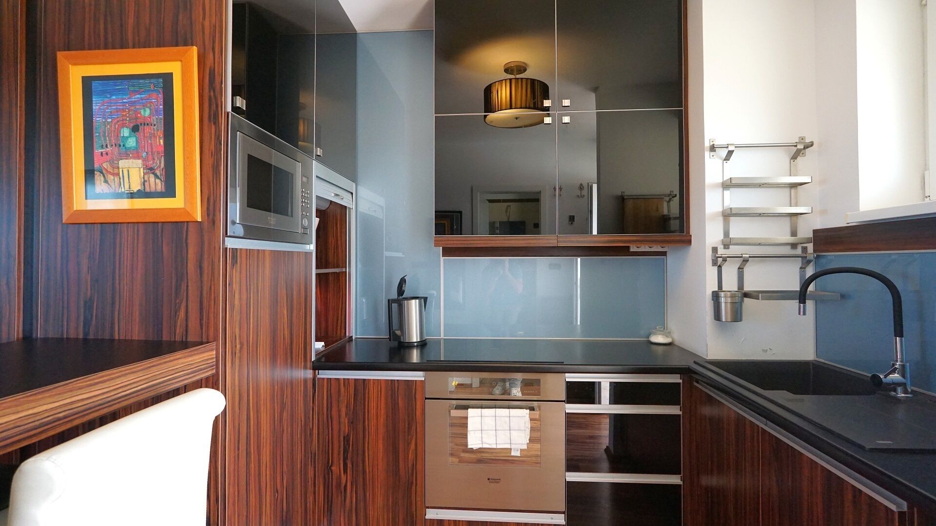 A compact kitchen with a microwave, oven, and sink. There's a wooden cabinet and a framed picture on the wall.