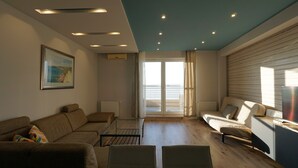 A modern living room with a sofa, a coffee table, and a TV. There is a large window with curtains and a view of the outside. - Club218 Wellness Apartments (Siófok)