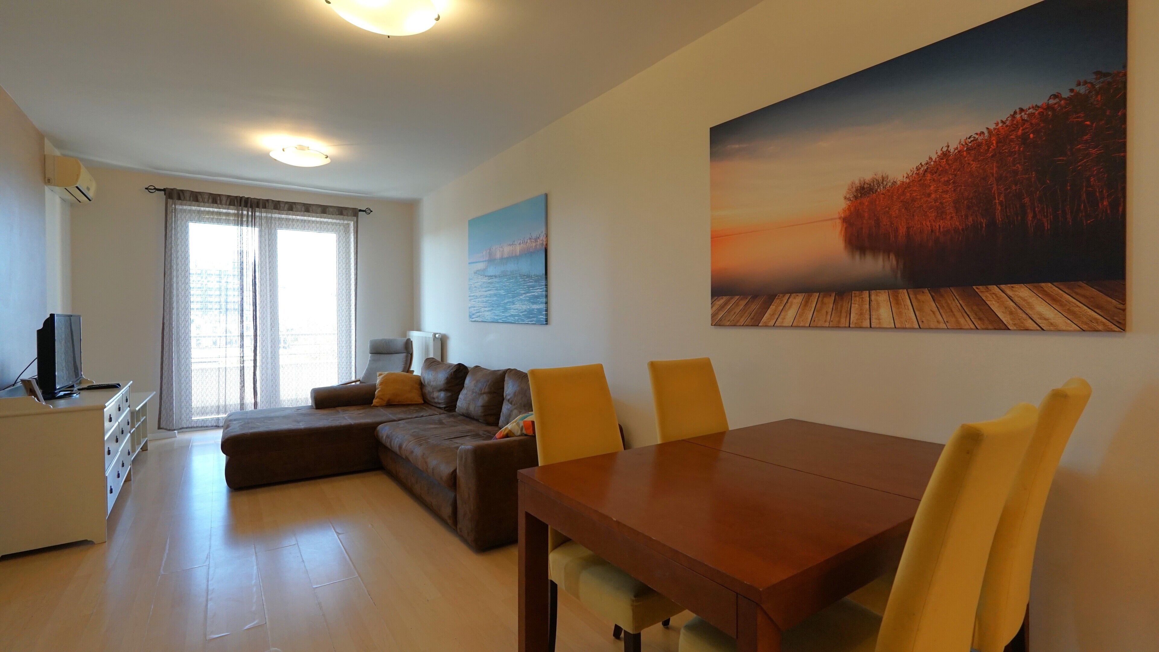 A modern living room with a sofa, dining table, and chairs. There is a painting on the wall and a window with curtains.