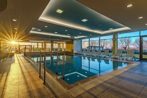 A swimming pool with a sunlit interior, clear glass walls, and a view of the outdoors. - Club218 Wellness Apartments (Siófok)
