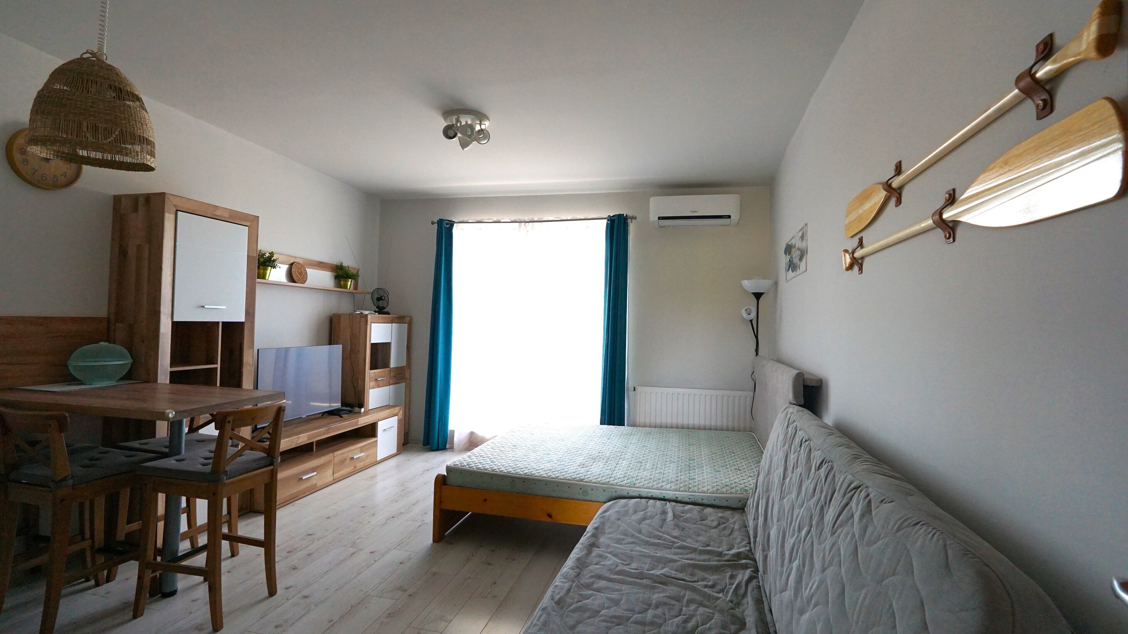 A small, well-lit room with a bed, a desk, a chair, a hanging light fixture, and a window with curtains.