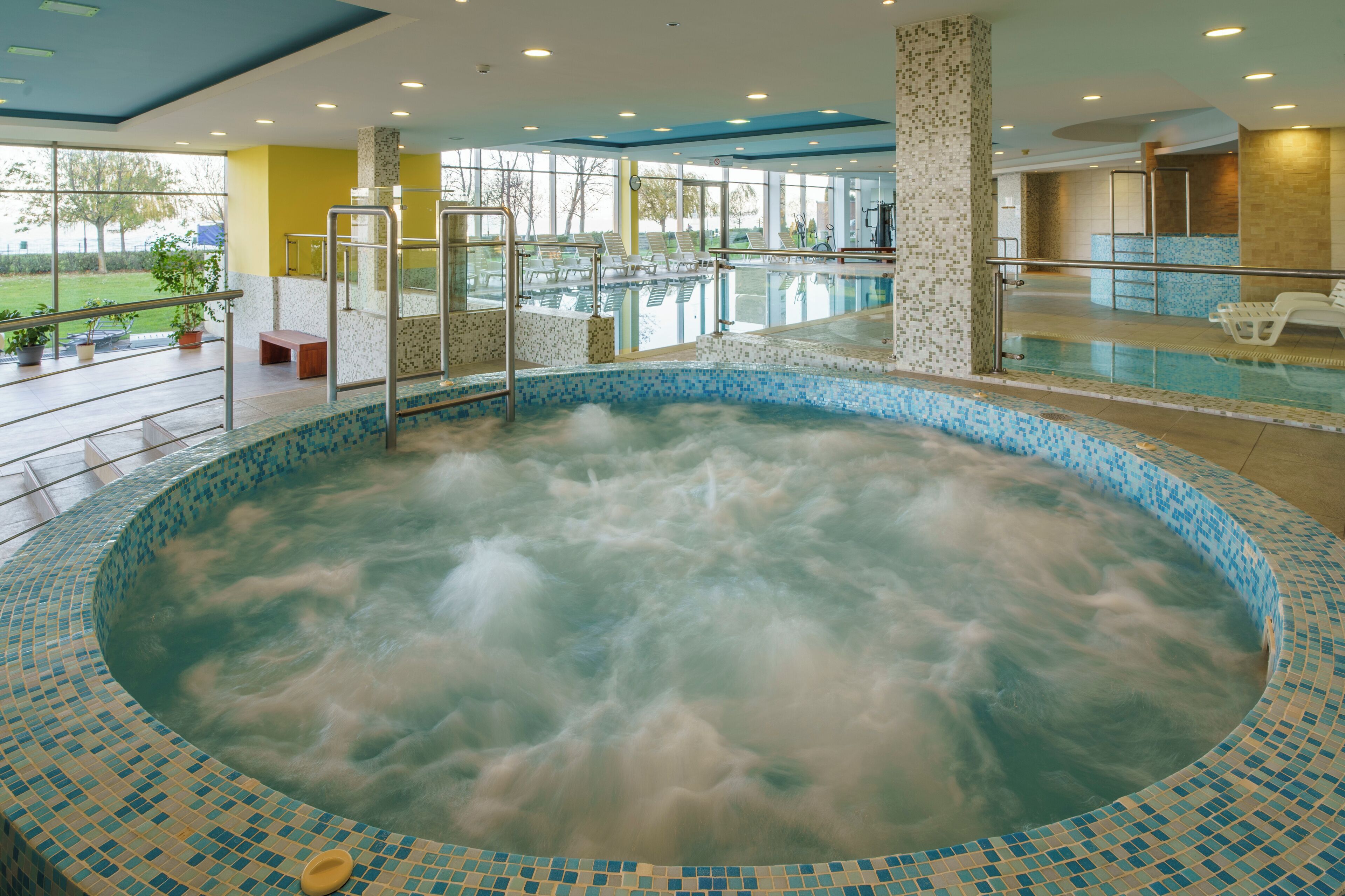 Vasca idromassaggio con bordo in mosaico, area piscina interna con poltrone relax e vista all'esterno tramite ampie finestre.
