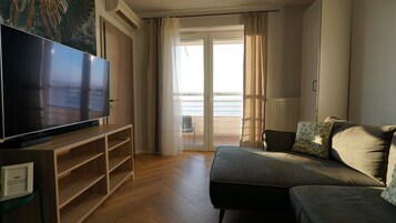 A modern living room with a flat-screen TV on a wooden stand, a sofa with cushions, and a sliding glass door leading to a balcony.