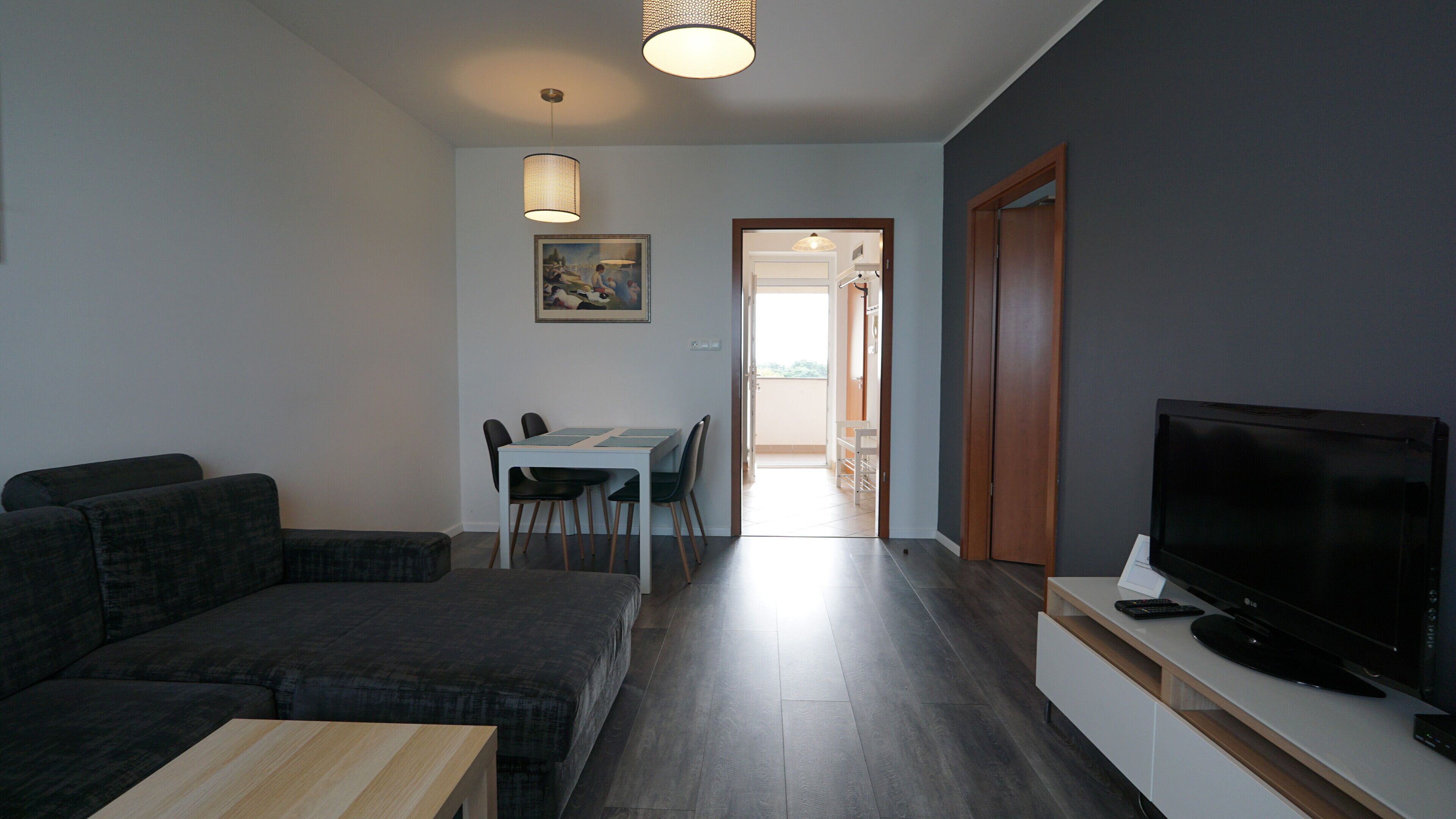 Una sala de estar con un sofá de color oscuro, una mesa de centro de madera y un televisor de pantalla plana sobre un mueble blanco. Hay un comedor con una mesa y sillas, y un cuadro en la pared.