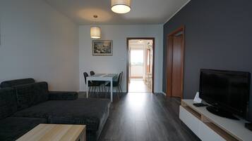 Una sala de estar con un sofá de color oscuro, una mesa de centro de madera y un televisor de pantalla plana sobre un mueble blanco. Hay un comedor con una mesa y sillas, y un cuadro en la pared.