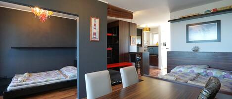 A modern hotel room with two beds, a dining table, and a kitchen area.
