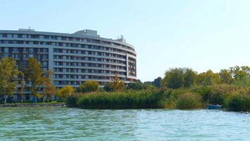 A large, multi-story hotel with a curved facade, located near a body of water with trees and a small boat visible.
