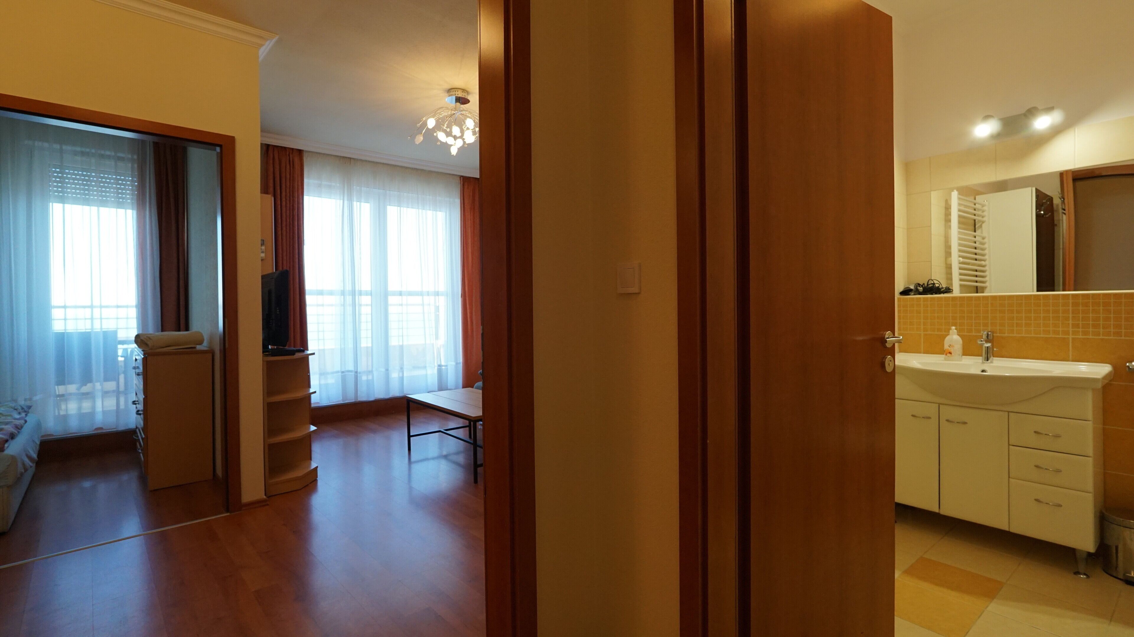 Una habitación de hotel moderna con piso de madera, zona de comedor, sala de estar con sofá y baño con lavamanos y espejo.