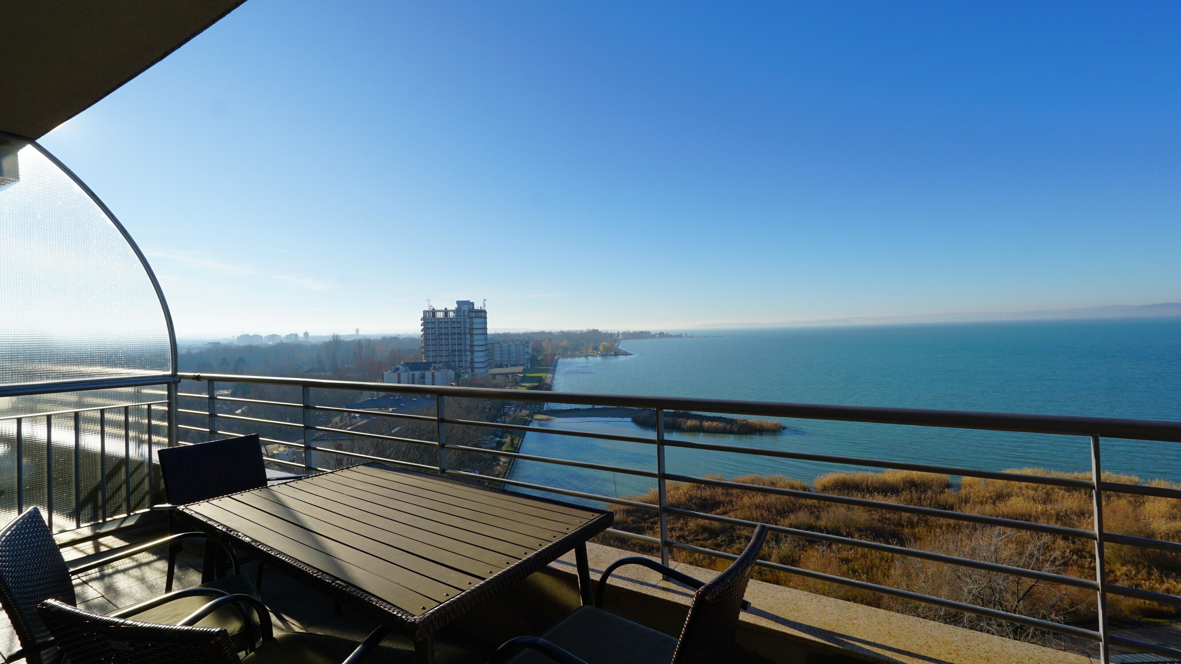 Un balcone con tavolo e sedie, affacciato su uno specchio d'acqua e un panorama urbano in lontananza.