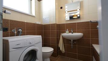 Un baño pequeño con lavadora de carga frontal, inodoro y lavamanos con espejo superior.
