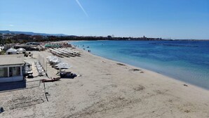 A beach with a row of sunbeds and umbrellas, a building with a dome, and a clear blue sky. - Rina Sea Apartments (Alghero)