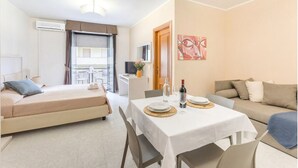 A hotel room with a bed, a dining table with plates and glasses, a sofa, a TV, and a balcony with a table and chairs. - Rina Sea Apartments (Alghero)