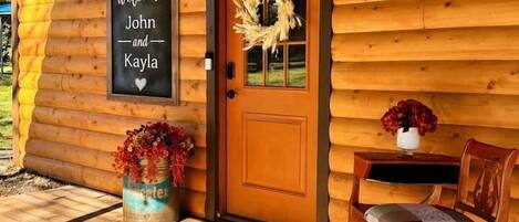 A wooden cabin with a welcome sign, a door with a wreath, a bench, and a planter with flowers.