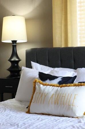 A bed with white bedding, a dark grey upholstered headboard, a lamp on a bedside table, and a pillow with 'STAY AWAY' embroidered on it.