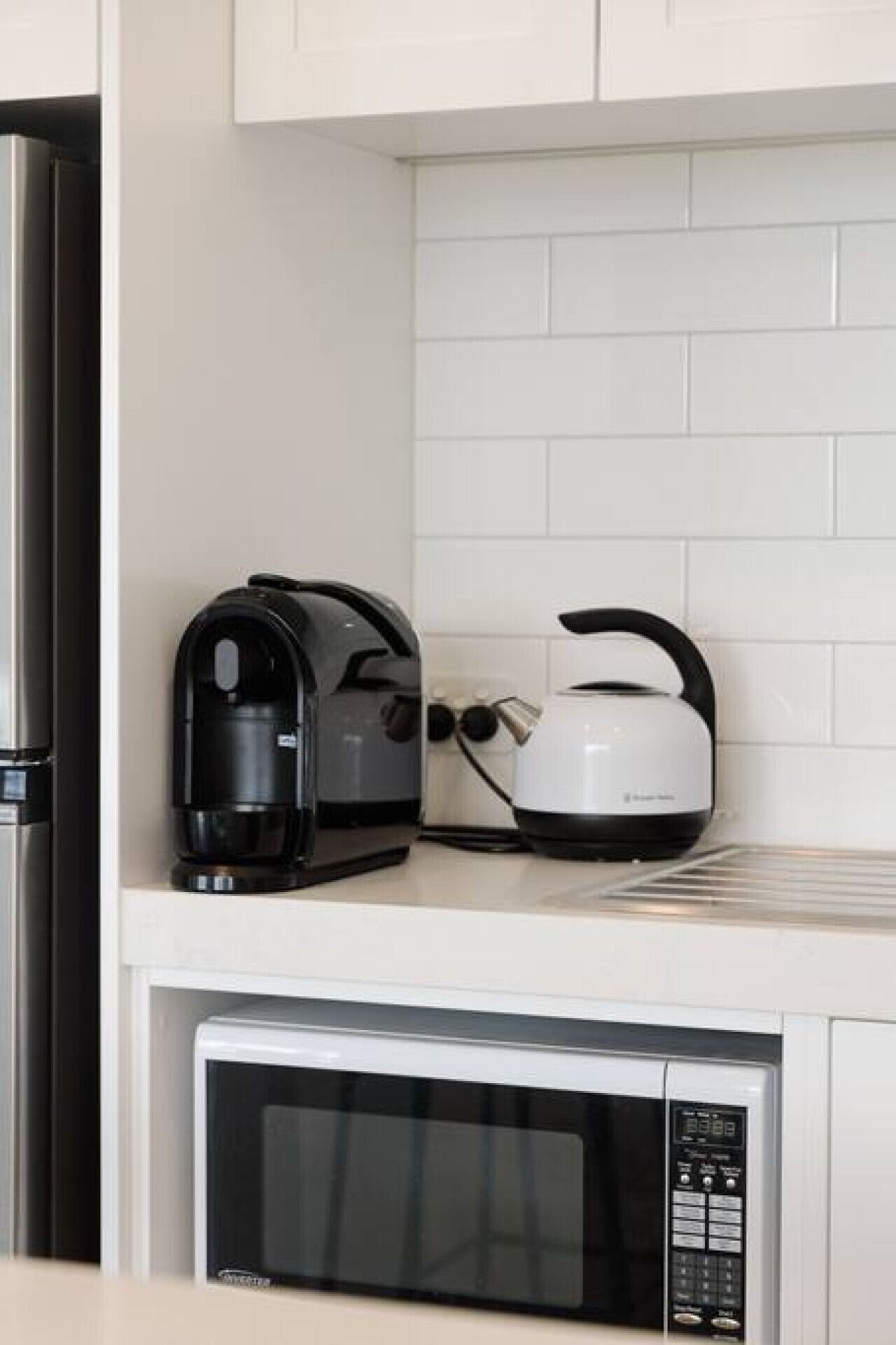 A kitchen with a stainless steel refrigerator, a black coffee maker, a white kettle, and a black microwave.