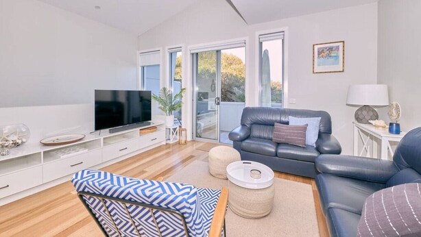 A modern living room with a flat-screen TV, leather sofas, and a large window offering a view of the outdoors. - Grove Hideaway - immaculate 2 bed townhouse (Ocean Grove)