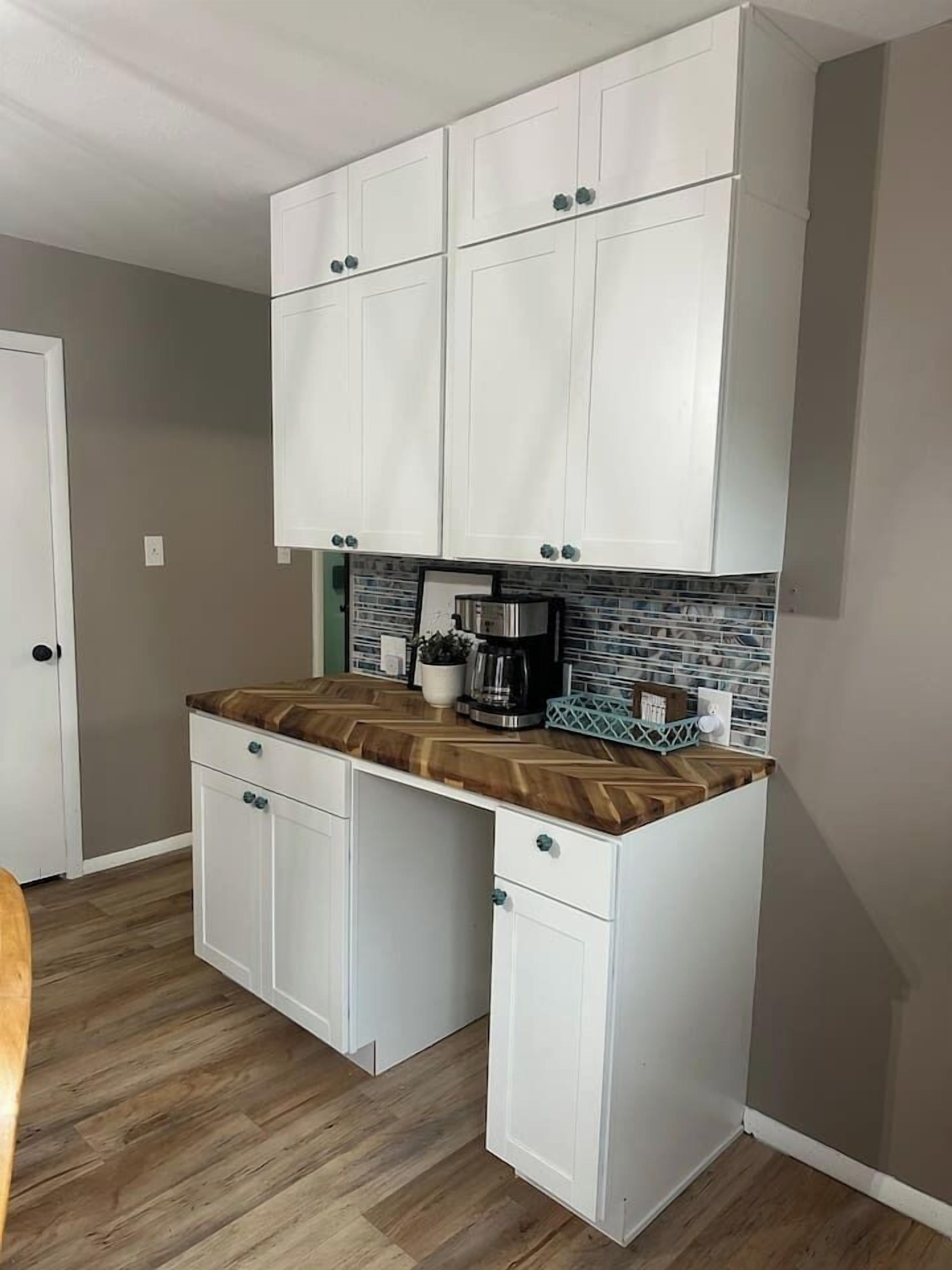 Cocina compacta con armarios blancos, encimera con motivo de espiga y cafetera.