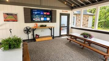 Una habitación con televisión, ventilador, banco y ventana con vista a follaje.