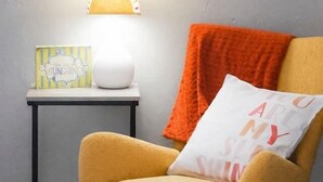 Een kamer met een schommelstoel, een bijzettafeltje met een lamp, en een wandplank met decoratieve spullen.