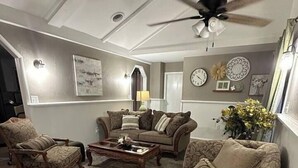 Een woonkamer met een bank, fauteuil en salontafel. Er is een plafondventilator en wanddecoraties.