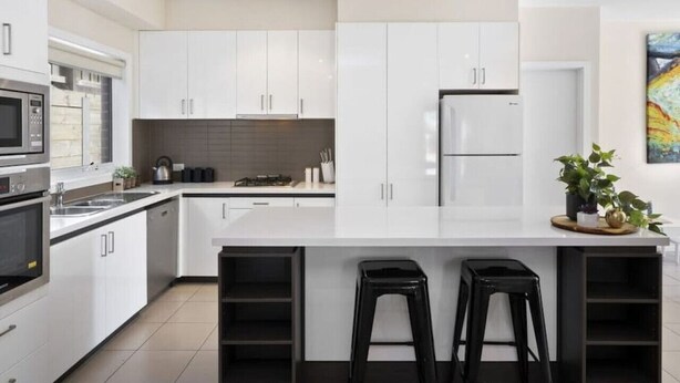 A modern kitchen with white cabinets, a central island with stools, stainless steel appliances, and a potted plant. - Oceans 10 (Ocean Grove)