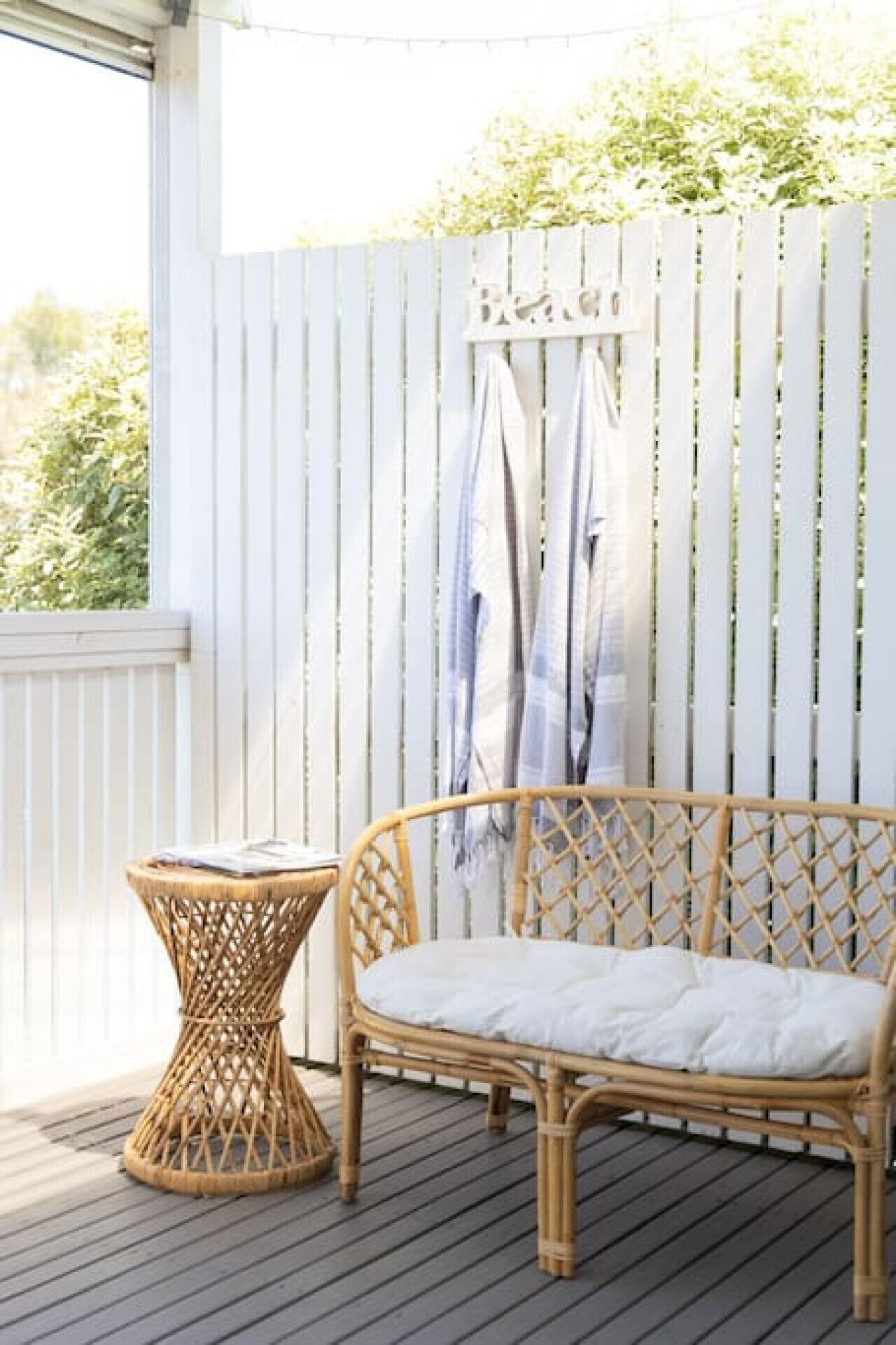 A wicker sofa with a white cushion on a wooden deck, a small wicker table, and a white fence with vertical slats.