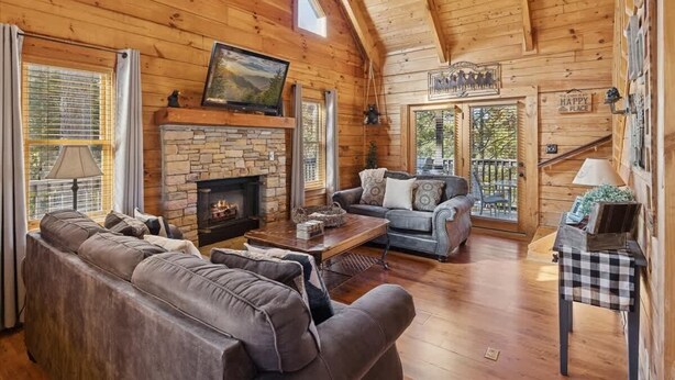 A cozy log cabin interior with a stone fireplace, leather sofas, and a large window overlooking a deck. - Firefly Holler by Beyond Expectations: Dog Friendly 3BR Cabin w/ EV Charger (Pigeon Forge)