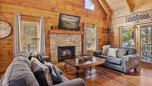 A cozy log cabin interior with a stone fireplace, a large window, and a comfortable seating area. - Firefly Holler by Beyond Expectations: Dog Friendly 3BR Cabin w/ EV Charger (Pigeon Forge)