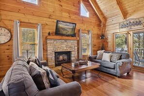 A cozy log cabin interior with a stone fireplace, a large window, and a comfortable seating area. - Firefly Holler by Beyond Expectations: Dog Friendly 3BR Cabin w/ EV Charger (Pigeon Forge)