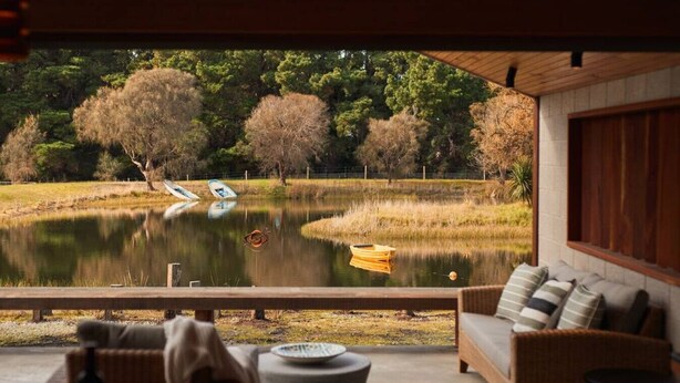 A view from a room with a large window overlooking a lake, a wooden deck, and a wicker sofa.