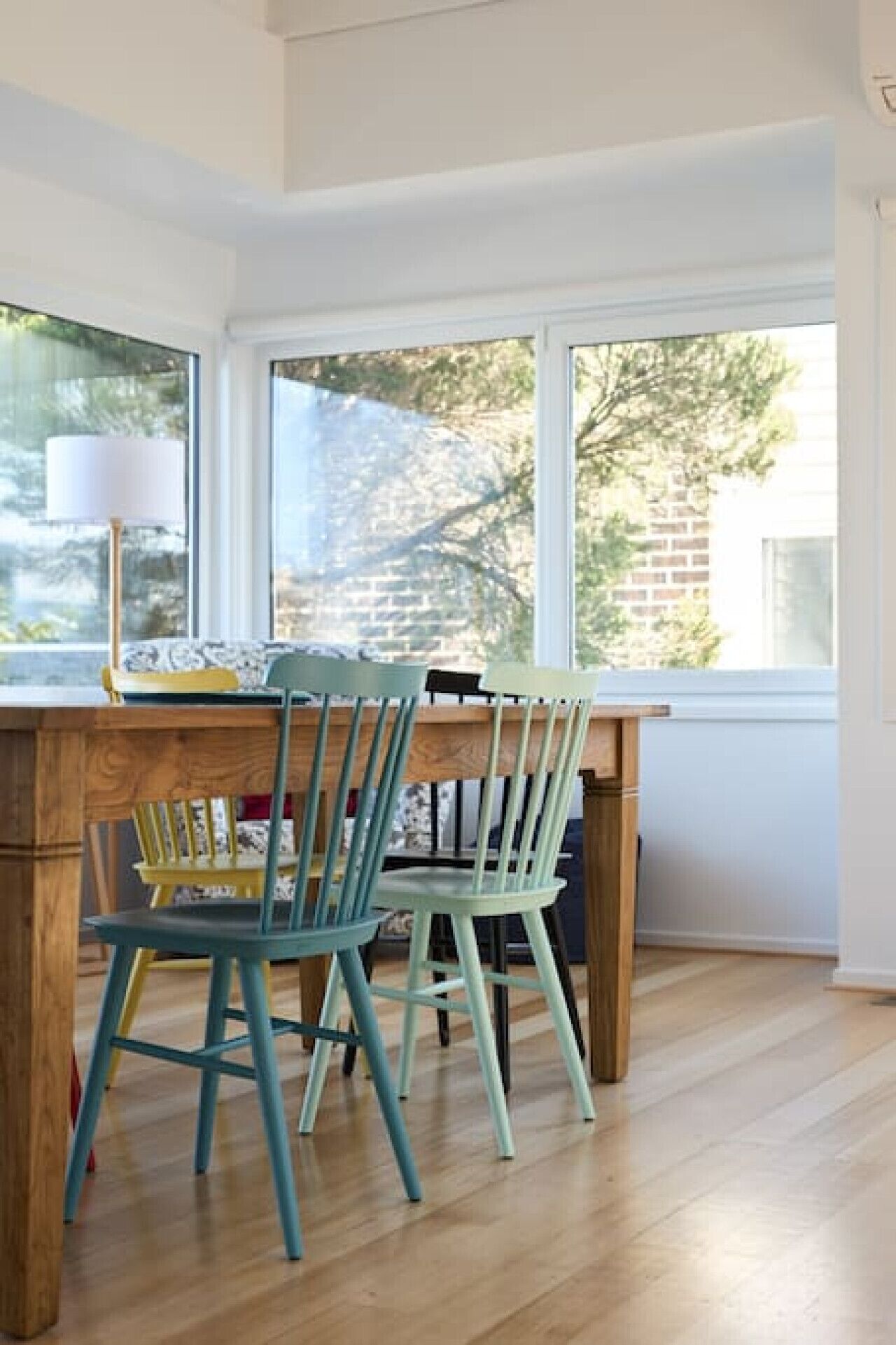 A dining area with a wooden table and two chairs, a lamp, and a view of trees and a brick wall through large windows.
