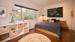 Un dormitorio con un ventanal grande, una cama con colcha gris, una mesita de noche de madera, una lámpara de pie y un cuadro colgado en la pared.