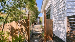 Un sendero estrecho que conduce a una casa, con una cerca de madera a un lado y un muro exterior blanco al otro.