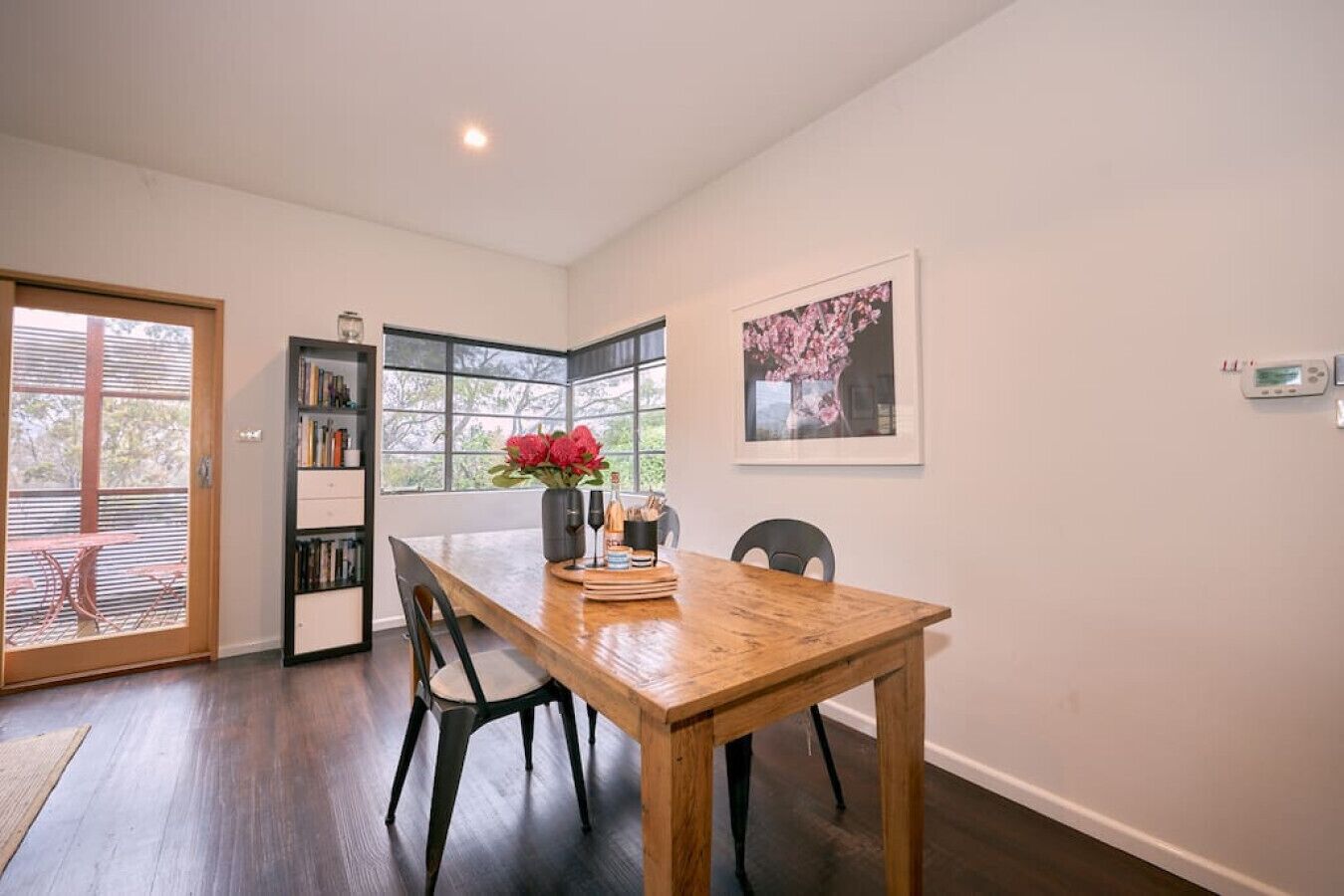 A dining area with a wooden table, chairs, a bookshelf, and a window with a view.