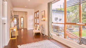 Una habitación amplia con un gran ventanal, piso de madera, una silla blanca y una pequeña mesa con una lámpara.